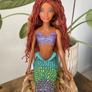 May include: A mermaid doll with long, auburn braids, a sparkling purple top, and a color-shifting tail. The tail transitions from teal to pink, with a light blue accent at the waist. A star-shaped charm hangs above the doll.