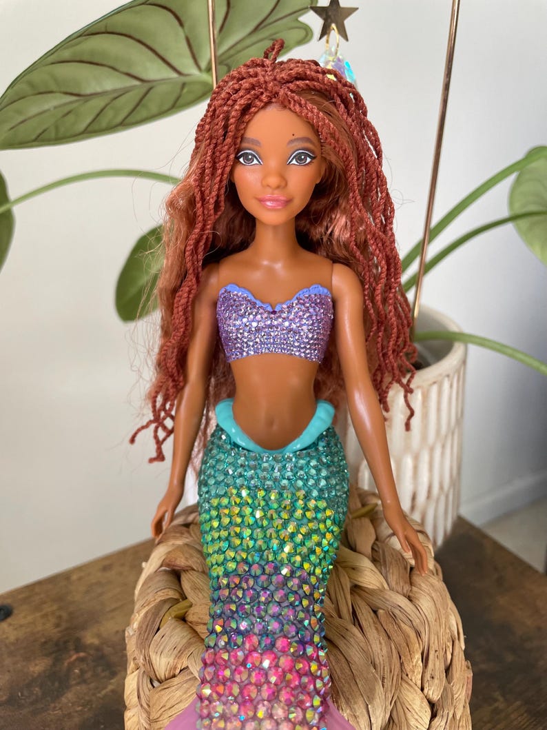May include: A mermaid doll with long, auburn braids, a sparkling purple top, and a color-shifting tail. The tail transitions from teal to pink, with a light blue accent at the waist. A star-shaped charm hangs above the doll.