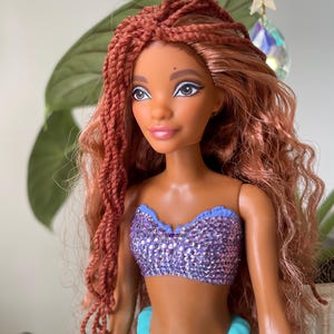 May include: A doll with brown skin, red braided hair, and a sparkling purple top. The doll has a turquoise tail and is decorated with a star and crystal. The doll is a mermaid.