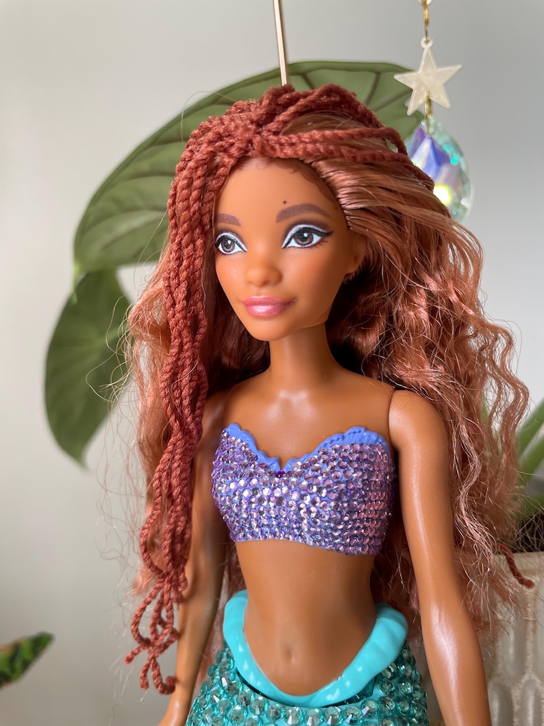 May include: A doll with brown skin, red braided hair, and a sparkling purple top. The doll has a turquoise tail and is decorated with a star and crystal. The doll is a mermaid.