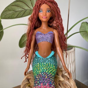 May include: A mermaid doll with auburn hair and a purple sequined top. The doll has a turquoise and iridescent sequined tail, and a pink fin. The doll is sitting on a woven basket.