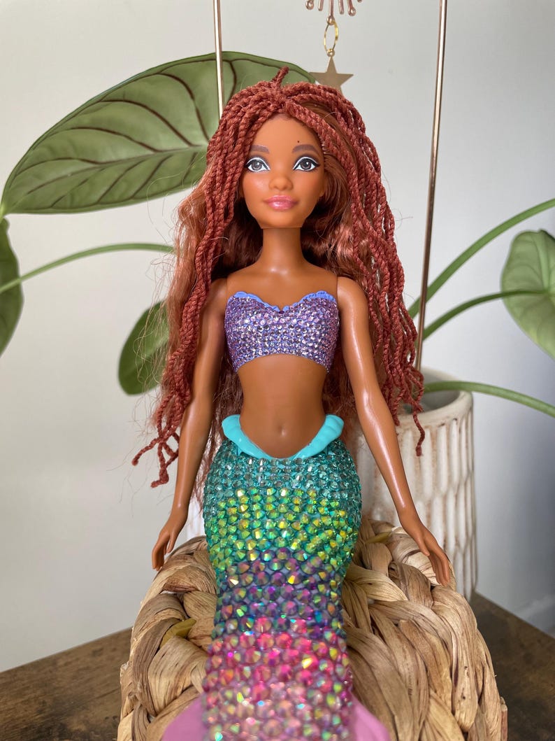 May include: A mermaid doll with auburn hair and a purple sequined top. The doll has a turquoise and iridescent sequined tail, and a pink fin. The doll is sitting on a woven basket.