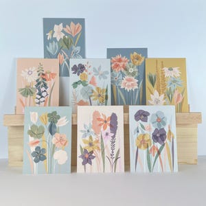 May include: A selection of floral art prints showcasing diverse flower arrangements in soft pastel hues. The prints are presented on a wooden stand against a light blue backdrop. Each print features unique botanical illustrations.