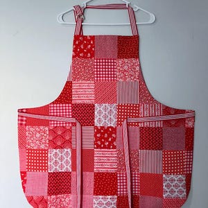 Pode incluir: Um avental de patchwork vermelho e branco pendurado em um cabide branco. O avental apresenta v&aacute;rios padr&otilde;es, incluindo bolinhas, listras e desenhos florais. Possui um babador, dois bolsos e al&ccedil;as listradas em vermelho e branco.