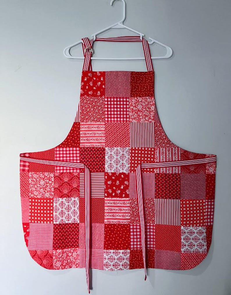 Pode incluir: Um avental de patchwork vermelho e branco pendurado em um cabide branco. O avental apresenta v&aacute;rios padr&otilde;es, incluindo bolinhas, listras e desenhos florais. Possui um babador, dois bolsos e al&ccedil;as listradas em vermelho e branco.