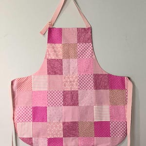 Pode incluir: Um avental de patchwork rosa com uma grade de retalhos de tecido quadrados. Os retalhos exibem padr&otilde;es como bolinhas, listras e desenhos florais. O avental tem uma al&ccedil;a de pesco&ccedil;o e la&ccedil;os na cintura, todos em tecidos rosa coordenados.