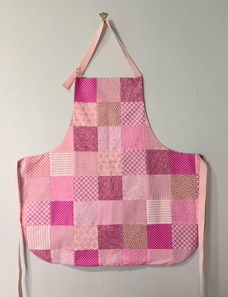 Pode incluir: Um avental de patchwork rosa com uma grade de retalhos de tecido quadrados. Os retalhos exibem padr&otilde;es como bolinhas, listras e desenhos florais. O avental tem uma al&ccedil;a de pesco&ccedil;o e la&ccedil;os na cintura, todos em tecidos rosa coordenados.