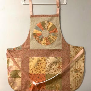 Pode incluir: Um avental acolchoado com um padr&atilde;o floral em tons de rosa, bege e verde. O avental apresenta um design circular no centro e la&ccedil;os de fita rosa. Pendurado num cabide branco.
