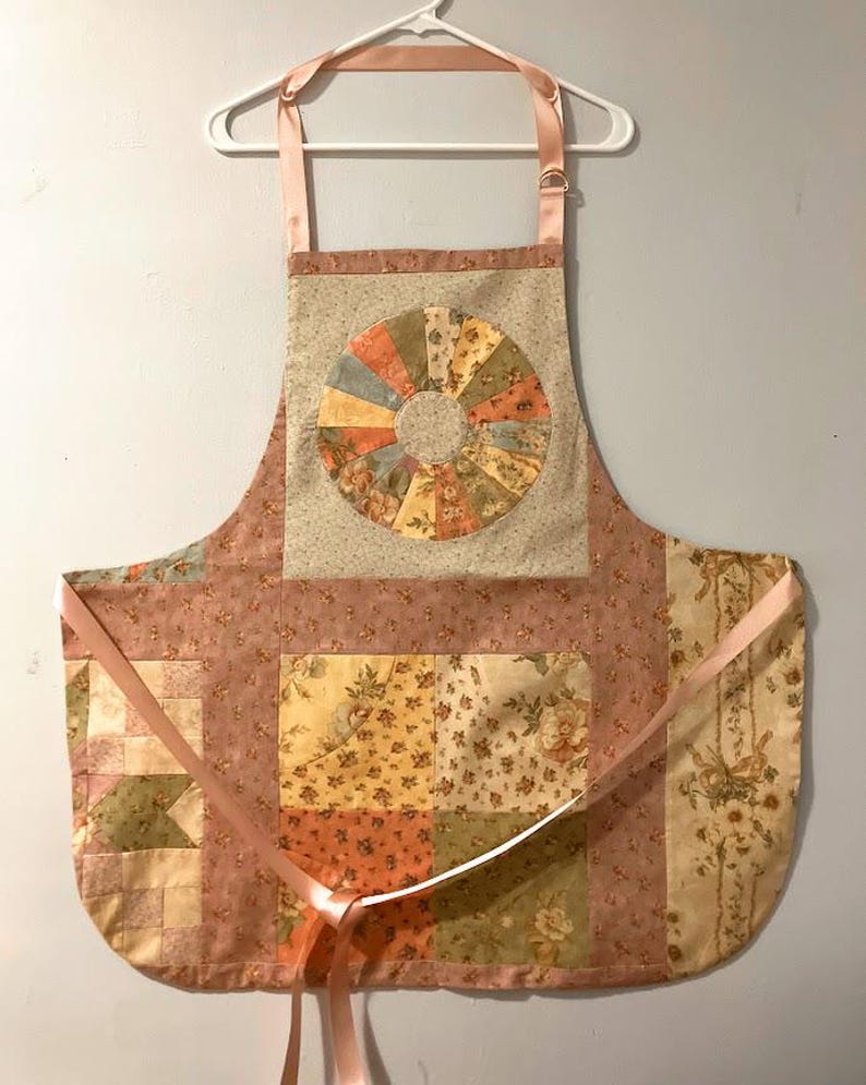 Pode incluir: Um avental acolchoado com um padr&atilde;o floral em tons de rosa, bege e verde. O avental apresenta um design circular no centro e la&ccedil;os de fita rosa. Pendurado num cabide branco.