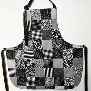 Pode incluir: Um avental de patchwork com uma al&ccedil;a de pesco&ccedil;o preta e la&ccedil;os na cintura. O avental apresenta uma variedade de quadrados estampados em preto, branco e cinza, incluindo bolinhas, listras e desenhos florais. O avental est&aacute; pendurado em um fundo branco.