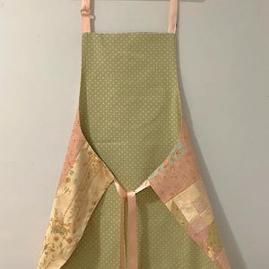 Pode incluir: Avental feito &agrave; m&atilde;o com um padr&atilde;o de bolinhas verde claro. Pain&eacute;is de patchwork floral em tons de rosa, creme e verde adornam as laterais. Possui uma fita e al&ccedil;as rosa, pendurado em um cabide branco.