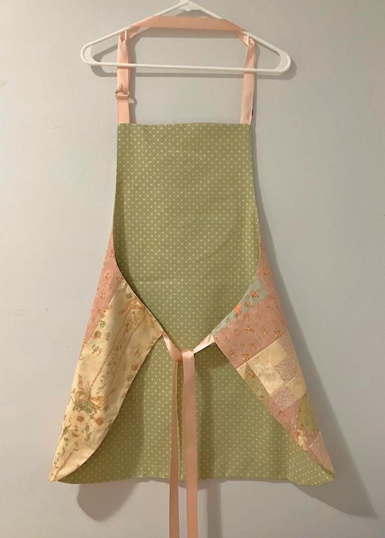 Pode incluir: Avental feito &agrave; m&atilde;o com um padr&atilde;o de bolinhas verde claro. Pain&eacute;is de patchwork floral em tons de rosa, creme e verde adornam as laterais. Possui uma fita e al&ccedil;as rosa, pendurado em um cabide branco.