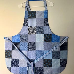 Pode incluir: Um avental patchwork azul com v&aacute;rios padr&otilde;es quadrados, incluindo florais e xadrez. Possui al&ccedil;as para o pesco&ccedil;o e cintura listradas. O avental &eacute; exibido em um cabide branco, mostrando seu design.