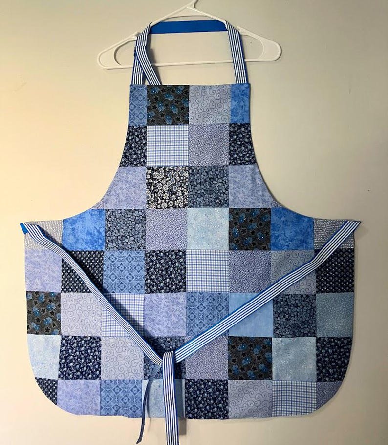 Pode incluir: Um avental patchwork azul com v&aacute;rios padr&otilde;es quadrados, incluindo florais e xadrez. Possui al&ccedil;as para o pesco&ccedil;o e cintura listradas. O avental &eacute; exibido em um cabide branco, mostrando seu design.
