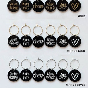 May include: Six black wine glass charms with different sayings in white and gold lettering. The charms are on a white background and are arranged in four rows of six. The sayings on the charms are "Sip Sip Hooray!" "Wine not?" "Cheers" "Wine o'clock" "Yay" and a heart. The charms are available in three different finishes: silver, gold, and white and gold.