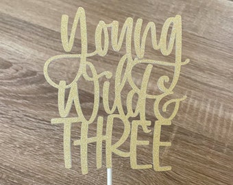 May include: Gold glitter cake topper with the text "Turning Wild & Three".