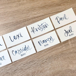 May include: A set of ten place cards with handwritten names in black ink on a cream background. The names are: Lexi, Cassidy, Joel, Nancy, Kristen, Paul, and two other names that are not fully visible.