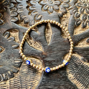 Puede incluir: Un brazalete de cuentas doradas con dos amuletos de ojo turco azules. El brazalete está sobre una superficie de madera marrón oscura con un diseño floral tallado.