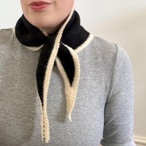 May include: A black and cream-colored knit scarf. The scarf is a triangular shape with a black center and cream-colored edges. The scarf is wrapped around the neck and tied in a knot.