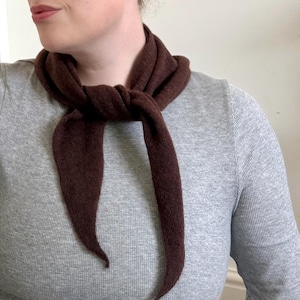 May include: A dark brown scarf tied around the neck. The scarf is made of a soft, textured material and is knotted at the front. The scarf's ends hang down, creating a simple yet elegant look. The person is wearing a gray long-sleeved shirt.