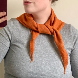 May include: An orange scarf is knotted around the neck. The scarf is made of a textured material and has two pointed ends. The person in the image is wearing a grey long-sleeved top.