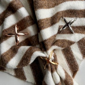 May include: Three metal scarf rings in gold, silver, and rose gold, each in a cross shape, are displayed on a brown and white striped knit scarf. The scarf is folded and draped, showcasing the rings' design and colour variations.