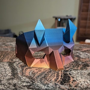 May include: A 3D printed mask with a geometric design. The mask is blue and orange with a gradient effect. The mask has two points on the top.