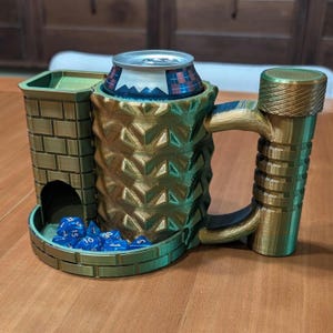 Op de afbeelding: Een groene en gouden 3D-geprinte mok met een ingebouwde dobbelsteentoren en een houder voor een blikje. De mok heeft een getextureerd ananasontwerp en een handvat in de vorm van een toren. Er zitten blauwe dobbelstenen in de toren.