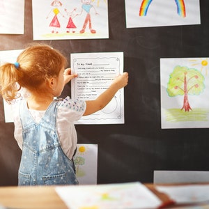May include: A young girl is standing in front of a chalkboard wall with several drawings on it. She is holding a piece of paper with the words "To my friend" written on it. The girl is wearing a white shirt and blue overalls.