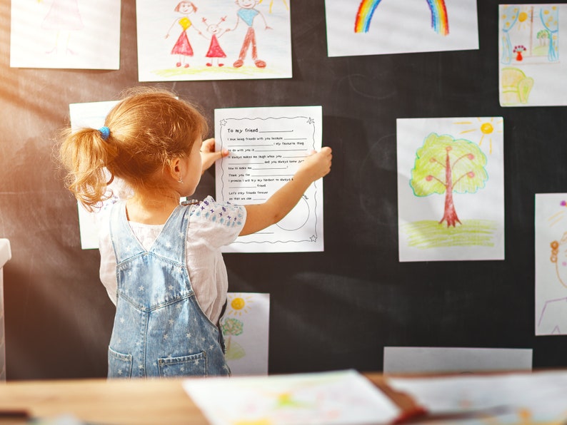 May include: A young girl is standing in front of a chalkboard wall with several drawings on it. She is holding a piece of paper with the words "To my friend" written on it. The girl is wearing a white shirt and blue overalls.