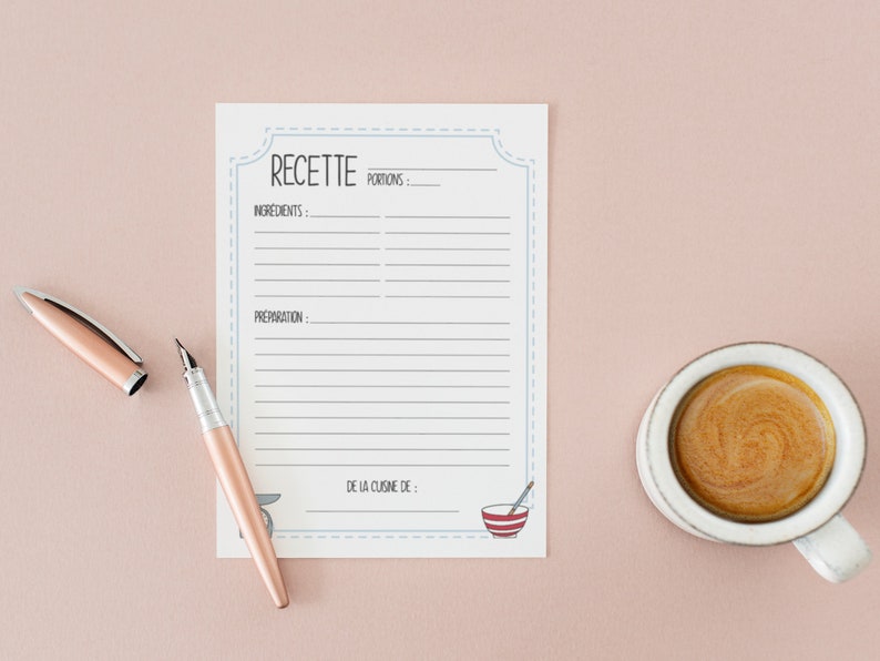 May include: A recipe notepad with a light blue border, the word "RECETTE" at the top, and lines for ingredients and preparation. A rose gold pen and a cup of coffee sit beside the notepad on a pink surface.
