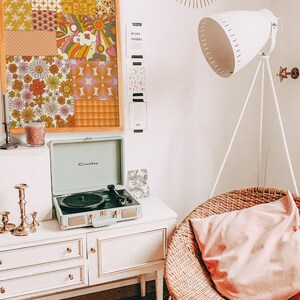 May include: A vintage-style record player with a green case and a black turntable sits on a white dresser with three drawers. A framed collage of colorful retro patterned fabric squares hangs on the wall behind the record player. A white floor lamp with a white shade sits in the corner of the room. A wicker chair with a pink cushion sits in the foreground.