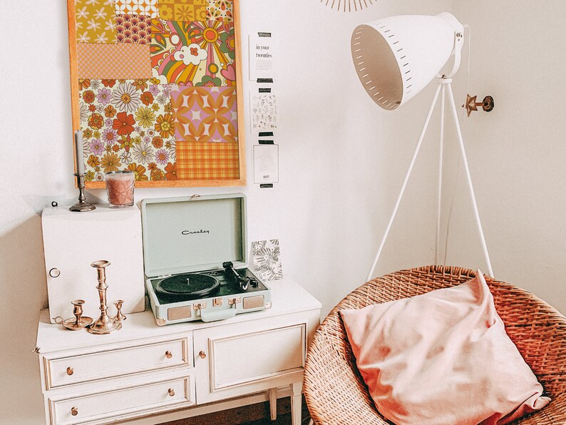 May include: A vintage-style record player with a green case and a black turntable sits on a white dresser with three drawers. A framed collage of colorful retro patterned fabric squares hangs on the wall behind the record player. A white floor lamp with a white shade sits in the corner of the room. A wicker chair with a pink cushion sits in the foreground.
