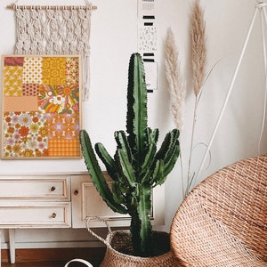 May include: A framed print with a patchwork design featuring various geometric and floral patterns in shades of orange, yellow, pink, and brown. The print is hanging on a white wall above a white dresser with three drawers.