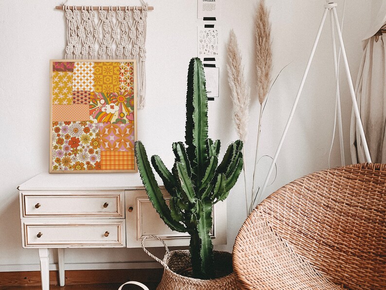 May include: A framed print with a patchwork design featuring various geometric and floral patterns in shades of orange, yellow, pink, and brown. The print is hanging on a white wall above a white dresser with three drawers.