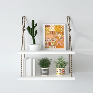 May include: A white two-tiered shelf with rope hangers, featuring a framed print of a colorful geometric pattern, a potted cactus, and two potted plants. The shelf is against a white wall.