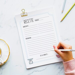 May include: A recipe notepad with the word "RECETTE" at the top, held by a gold binder clip. The notepad has lines for ingredients and preparation, with a scale and bowl illustration. A hand is writing on the notepad with a pen.