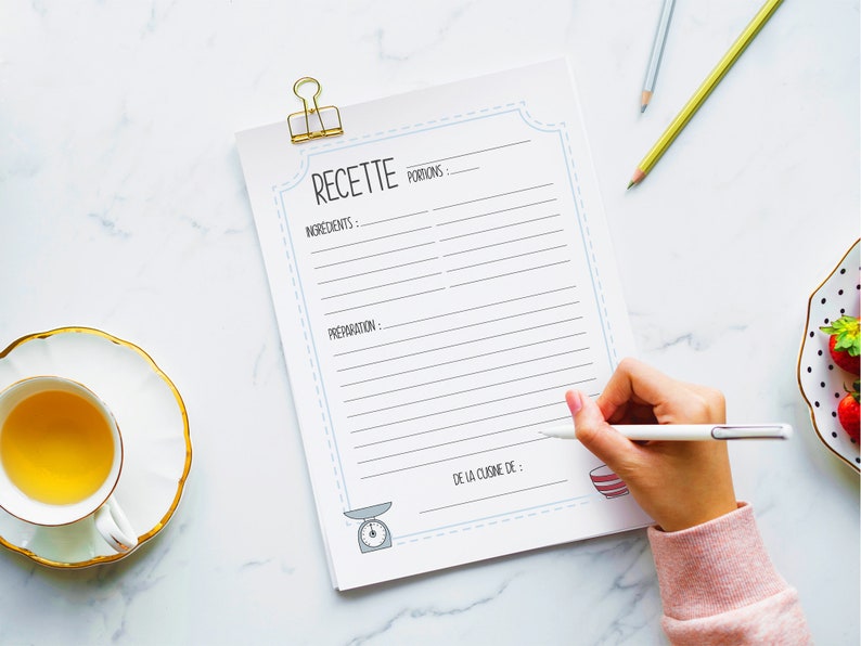 May include: A recipe notepad with the word "RECETTE" at the top, held by a gold binder clip. The notepad has lines for ingredients and preparation, with a scale and bowl illustration. A hand is writing on the notepad with a pen.