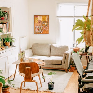 May include: A living room with a white couch, a wooden coffee table, and a large framed patchwork wall hanging with a floral pattern. There are plants in pots on the floor and shelves, and a hanging plant in a woven basket.