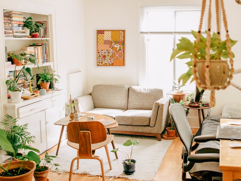 May include: A living room with a white couch, a wooden coffee table, and a large framed patchwork wall hanging with a floral pattern. There are plants in pots on the floor and shelves, and a hanging plant in a woven basket.
