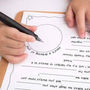 May include: A child's hand writes on a white sheet of paper with a black pen. The paper has a circle in the middle and text that says "Here's a drawing of you!" and "Let's stay friends forever so that we can... together... we're grown up!"