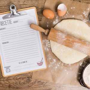 May include: A wooden rolling pin rests on rolled-out dough dusted with flour, alongside a recipe clipboard with the word "RECETTE" and a bowl of flour. Two eggs and measuring cups are also present, suggesting a baking theme.