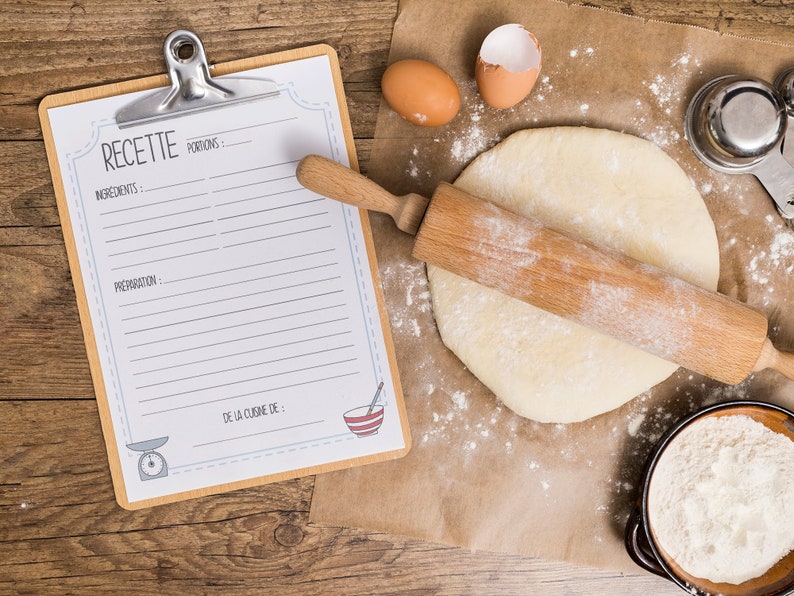 May include: A wooden rolling pin rests on rolled-out dough dusted with flour, alongside a recipe clipboard with the word "RECETTE" and a bowl of flour. Two eggs and measuring cups are also present, suggesting a baking theme.