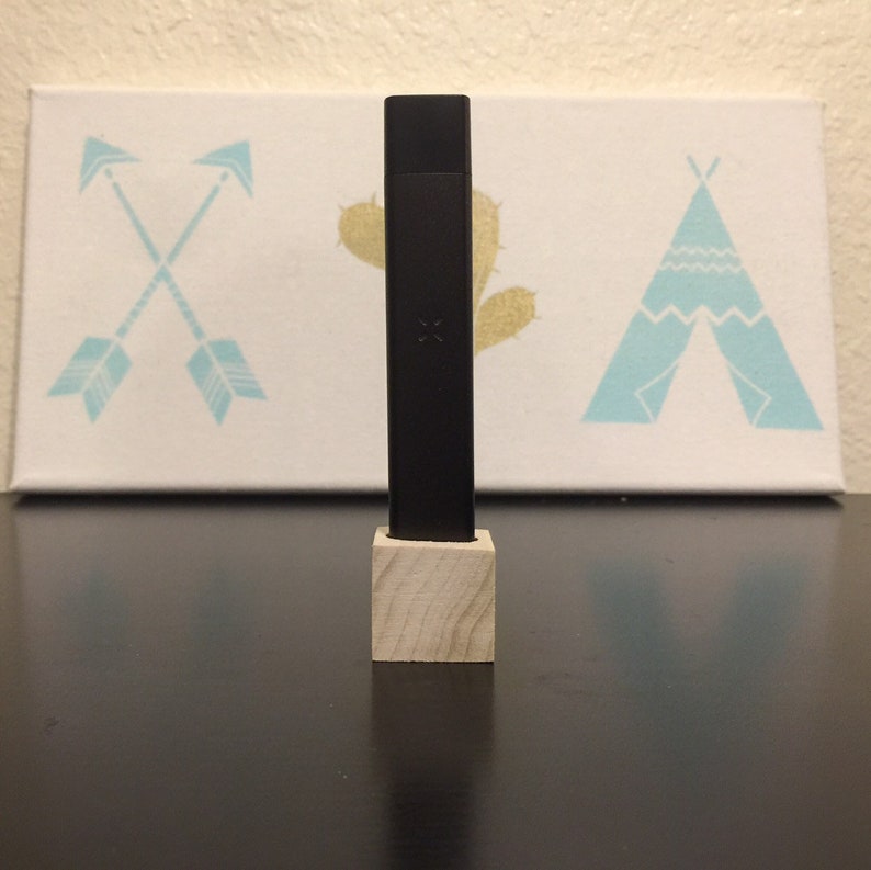 May include: A black rectangular vape pen with a wooden stand. The pen is sitting on a dark wooden surface in front of a white canvas with a blue teepee, two blue arrows, and a yellow cactus design.
