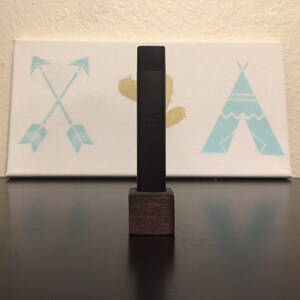 May include: A black vape pen with a wooden stand. The pen is standing on a dark surface in front of a white canvas with a blue and white teepee design.