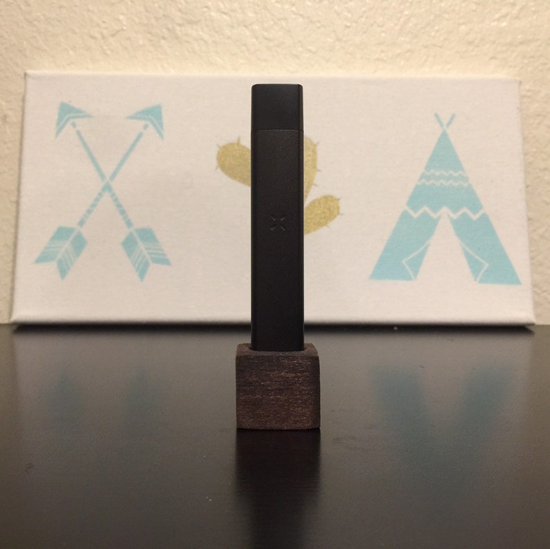 May include: A black vape pen with a wooden stand. The pen is standing on a dark surface in front of a white canvas with a blue and white teepee design.
