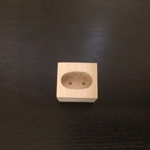 May include: A small wooden block with a cutout oval shape in the center. The oval has two small holes drilled into it.