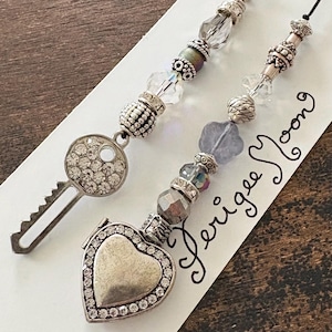 May include: Two beaded charms attached to a white card with the text Perigee Moon. One charm features a silver-toned key encrusted with clear rhinestones. The other charm features a silver-toned heart-shaped locket bordered by clear rhinestones.