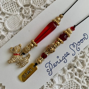 May include: Two red and gold beaded bookmarks with a small gold dog charm on one and the word "LOVE" on a tag on the other. The bookmarks are on a white card with the words "Perigee Moon" written in blue ink.