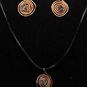 Può includere: Un set di collana e orecchini in filo di rame con una pietra preziosa trasparente e viola in ogni pezzo. La collana è su un cordoncino nero.